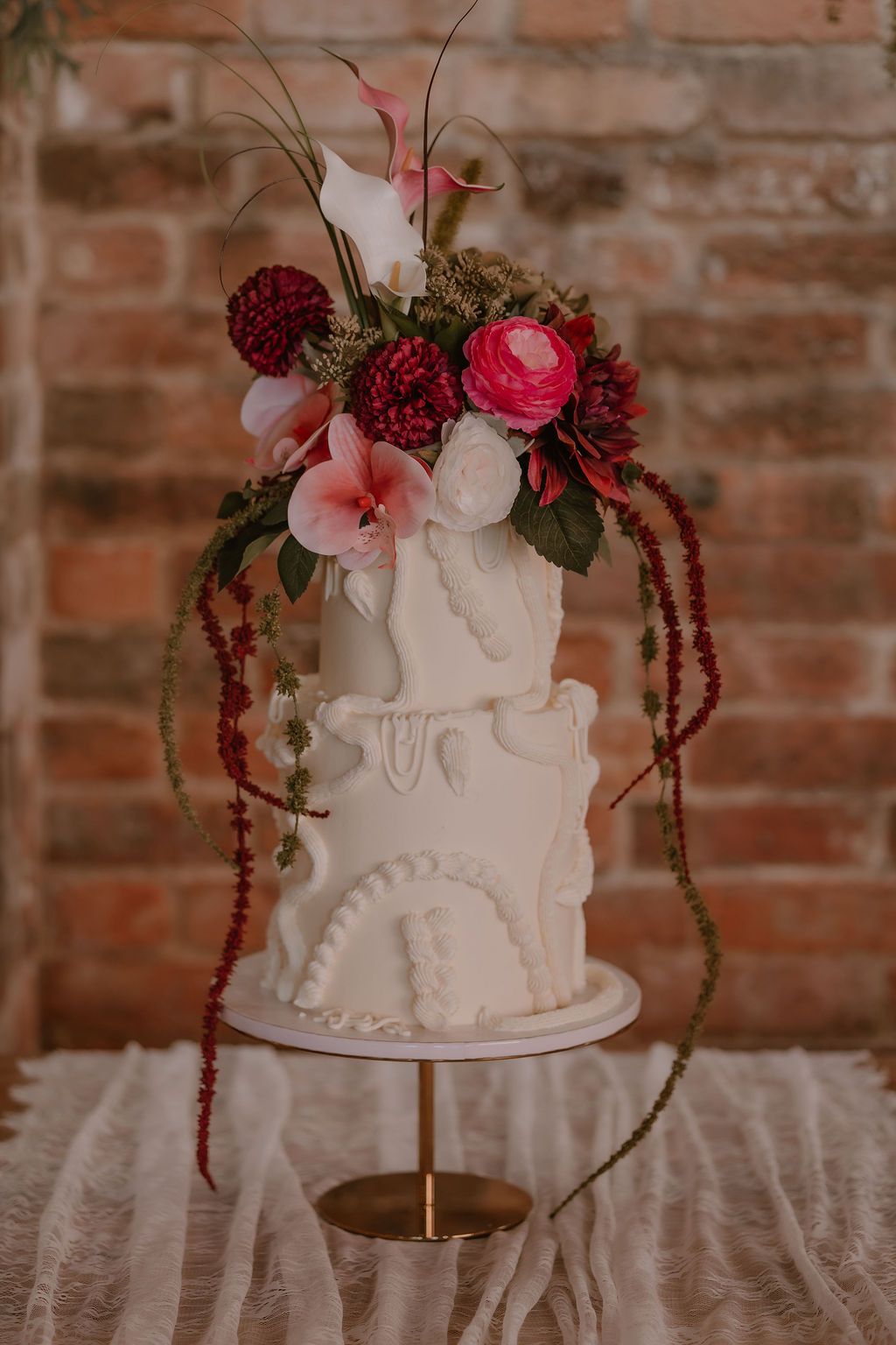 Two-tier white buttercream wedding cake with sculptural piping, topped with bold pink and burgundy florals against an exposed brick wall